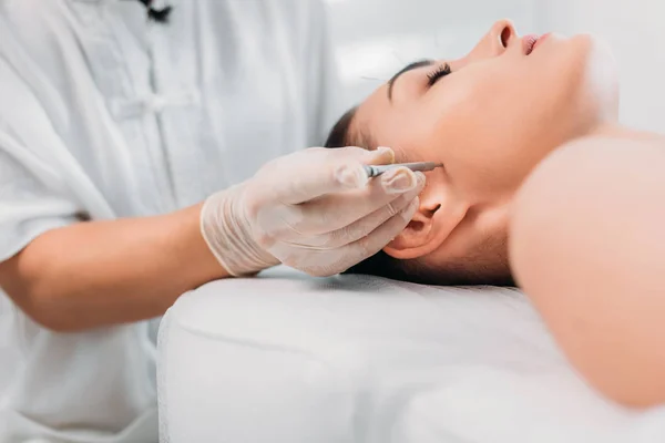 Vista parcial del cosmetólogo colocando aguja en la cara de las mujeres durante la terapia de acupuntura en el salón de spa - foto de stock