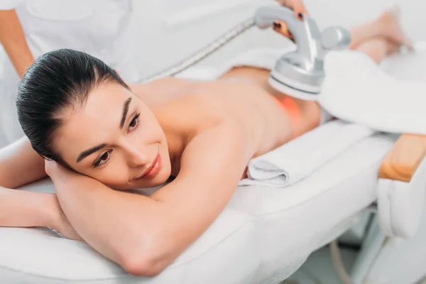 Hermosa mujer sonriente en la mesa de masaje recibiendo masaje eléctrico hecho por el cosmetólogo en el salón de spa - foto de stock