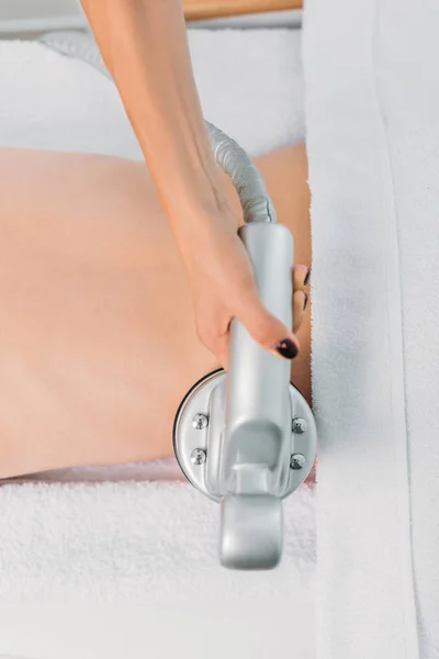 Vista parcial de la mujer en la mesa de masaje recibiendo masaje eléctrico realizado por el cosmetólogo en el salón de spa - foto de stock