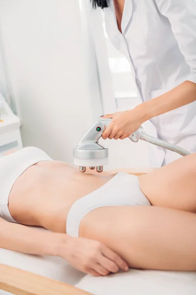 Cropped shot of cosmetologist making electrical massage on stomach to female client in white underwear in spa salon — Stock Photo