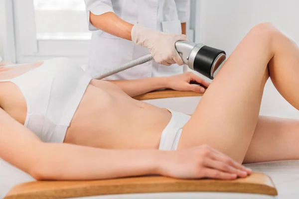 Частичный взгляд косметолога, делающего электрический массаж ноги женщине в белом нижнем белье в спа-салоне — стоковое фото