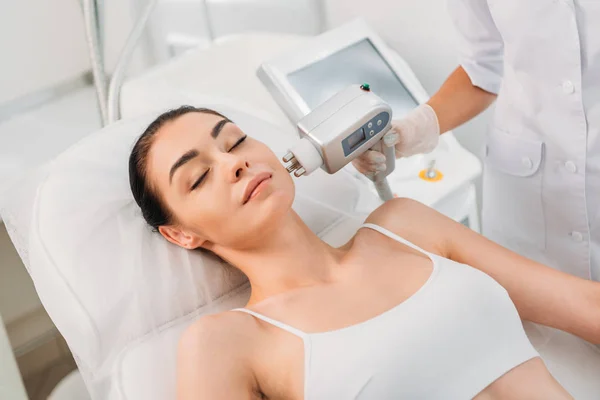 Vista parcial de la mujer recibiendo masaje eléctrico estimulante facial hecho por el cosmetólogo en el salón de spa - foto de stock