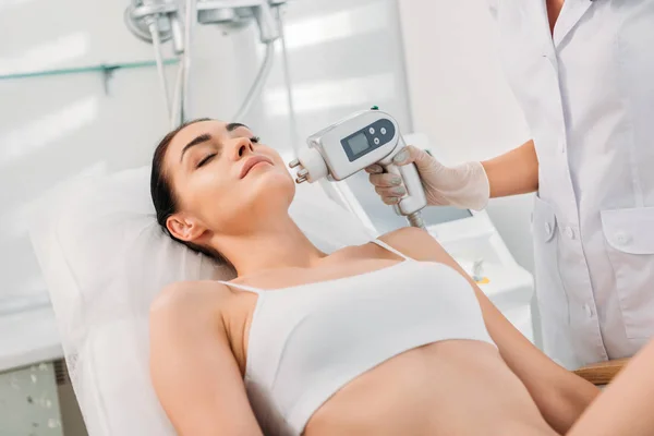 Vista parcial de la mujer en ropa interior recibiendo masaje eléctrico estimulante facial hecho por el cosmetólogo en el salón de spa - foto de stock