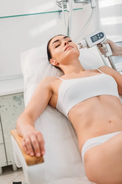 Vista parcial de la mujer en ropa interior recibiendo masaje eléctrico estimulante facial hecho por el cosmetólogo en el salón de spa - foto de stock
