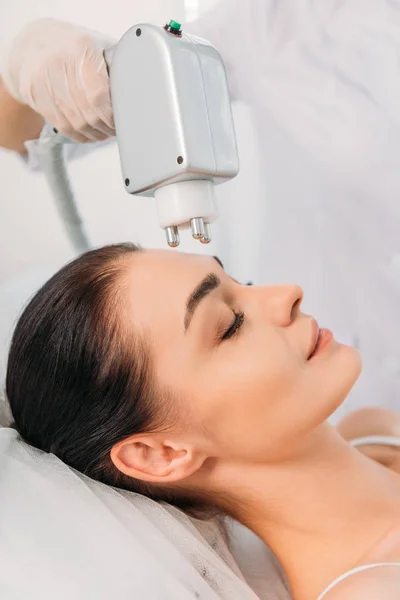 Partial view of woman getting facial microcurrent therapy made by cosmetologist in spa salon — Stock Photo