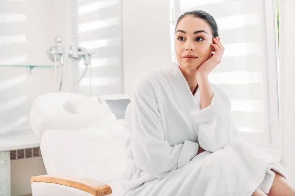 Bela mulher pensativa em roupão de banho branco sentado na mesa de massagem no salão de spa — Fotografia de Stock