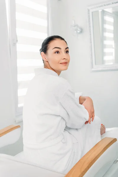 Hermosa mujer en albornoz blanco sentado en la mesa de masaje en el salón de spa - foto de stock