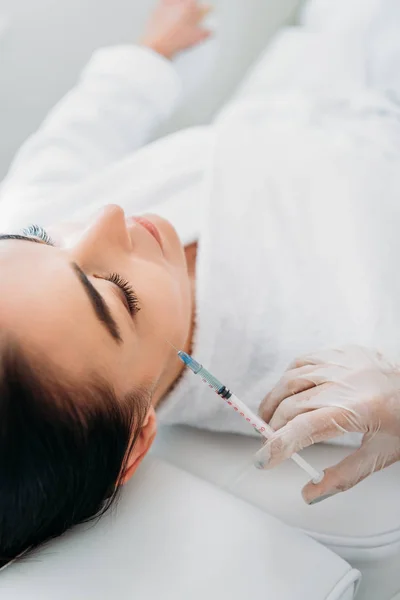 Inyección recortada de cosmetólogo en guante de látex con jeringa haciendo inyección de belleza a una hermosa mujer en el salón de spa - foto de stock