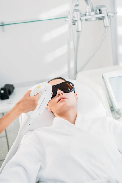 Mujer en gafas protectoras consiguiendo depilación láser hecha por cosmetólogo en salón de spa - foto de stock