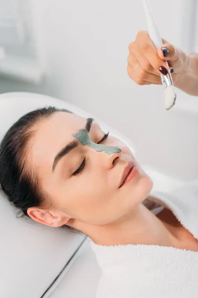 Vue partielle du cosmétologue appliquant un masque d'argile sur le visage féminin dans le salon de spa — Photo de stock