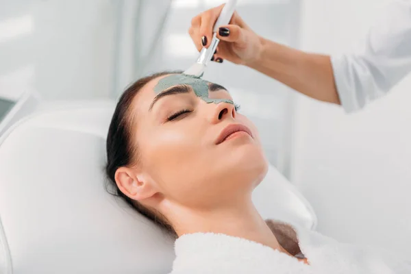 Vista parziale di cosmetologo che applica maschera di argilla con pennello su faccia femminile in salone di centro benessere — Foto stock