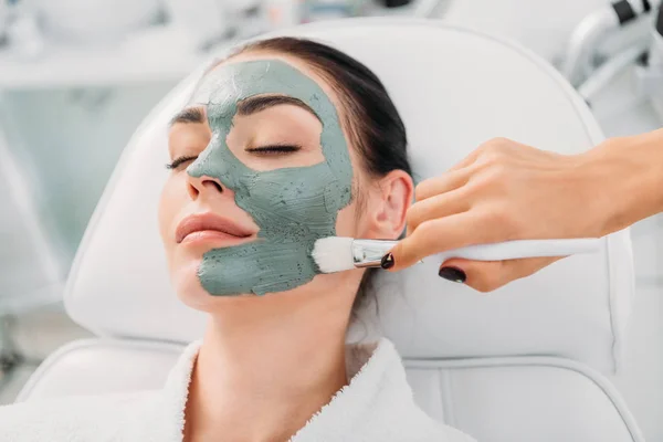 Vue partielle du cosmétologue appliquant un masque d'argile sur le visage féminin dans le salon de spa — Photo de stock