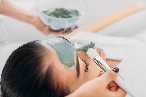 Vue partielle du cosmétologue appliquant un masque en argile avec pinceau sur le visage féminin dans le salon de spa — Photo de stock