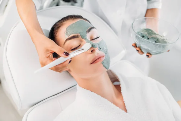 Teilansicht des Kosmetologen, der im Wellness-Salon Tonmaske mit Pinsel auf das weibliche Gesicht aufträgt — Stockfoto