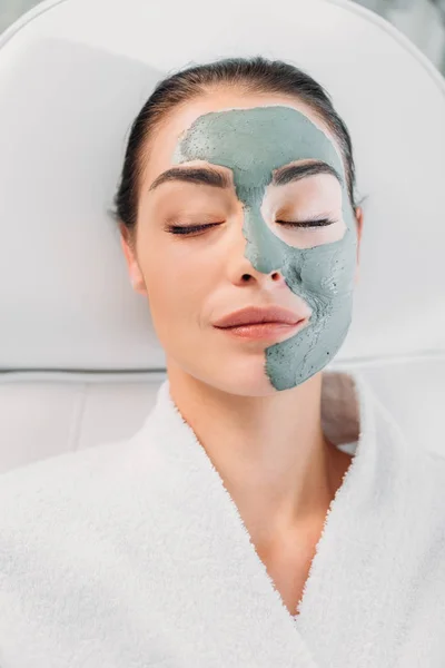 Overhead view of beautiful woman with eyes closed and clay mask on face in white bathrobe relaxing in spa salon — Stock Photo