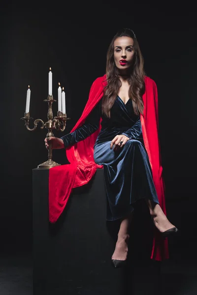 Beautiful vampire in red cloak licking teeth and holding antique candelabrum isolated on black — Stock Photo