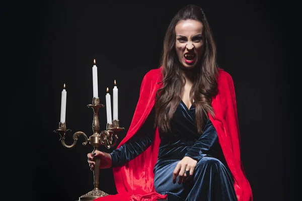 Woman in red cloak holding vintage candelabrum and showing vampire fangs isolated on black — Stock Photo