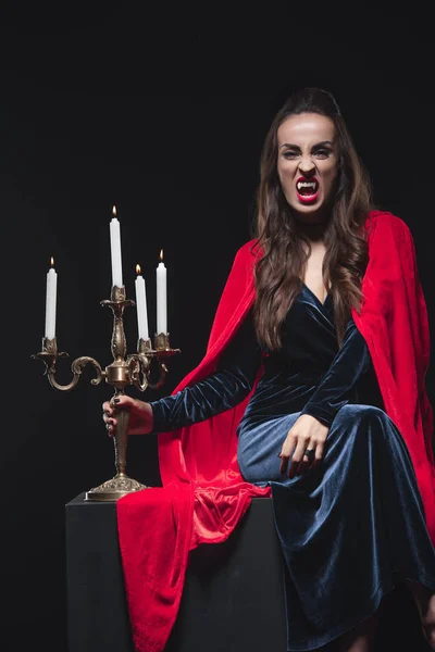 Dreadful woman in red cloak holding vintage candelabrum and showing vampire teeth isolated on black — Stock Photo