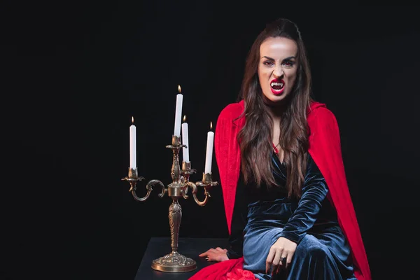 Femme sexy en costume de vampire assis près de vintage candelabrum isolé sur noir — Photo de stock