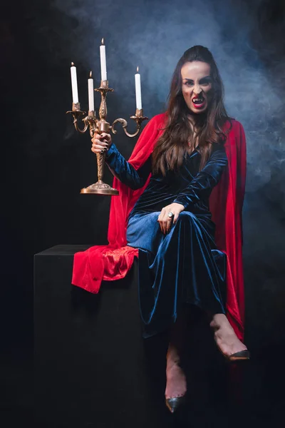 Beautiful woman in red cloak holding retro candelabrum and showing vampire teeth on dark background with smoke — Stock Photo