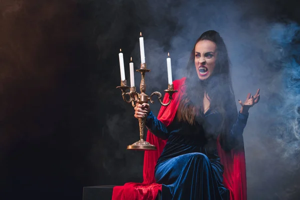 Mysteriöse Frau im Vampirkostüm mit antiken Kerzenleuchtern auf dunklem Hintergrund mit Rauch — Stockfoto