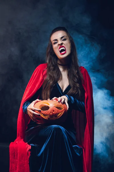 Woman in vampire costume holding jack o lantern on darkness with smoke — Stock Photo