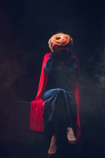 Woman in red cloak holding jack o lantern in front of face on dark background with smoke — Stock Photo