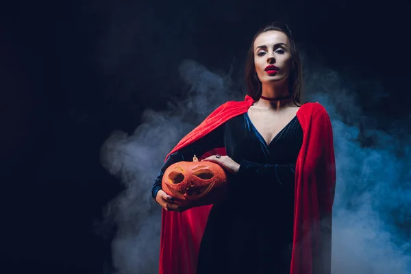 Woman in red cloak holding jack o lantern on dark background with smoke — Stock Photo