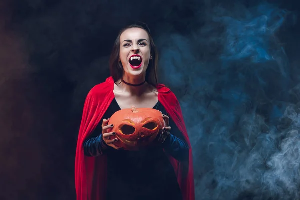 Mystic woman in vampire costume holding jack o lantern on dark background with smoke — Stock Photo