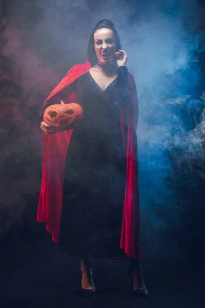 Attractive woman in vampire costume holding pumpkin on dark background with smoke — Stock Photo