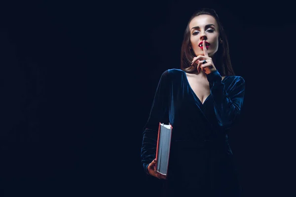 Vampire woman showing silence symbol and holding magic book isolated on black — Stock Photo