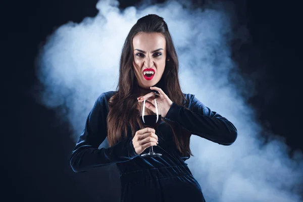 Grim vampire holding wineglass with blood on dark background with smoke — Stock Photo