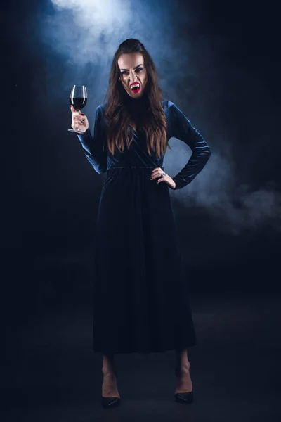 Grim vampire holding wineglass with blood on dark background with smoke — Stock Photo