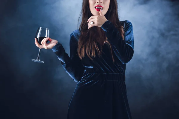 Vista cortada de vampiro segurando vinho com sangue e lambendo os dedos no fundo escuro com fumaça — Fotografia de Stock