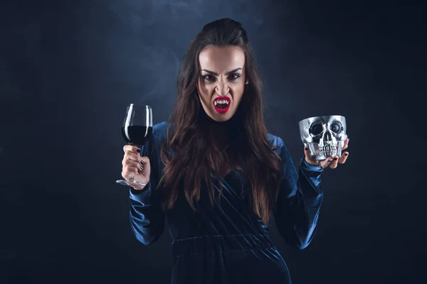 Vampire holding metal skull and wineglass with blood on darkness with smoke — Stock Photo