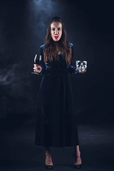 Vampire femme tenant crâne et verre à vin avec du sang sur fond sombre avec de la fumée — Photo de stock