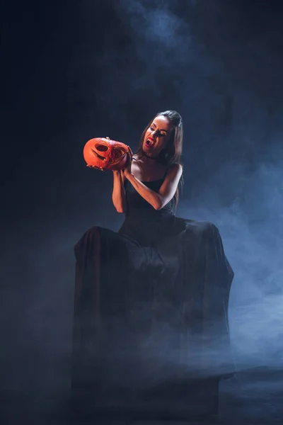 Espantosa mujer en traje de vampiro sosteniendo jack o linterna sobre fondo oscuro con humo - foto de stock