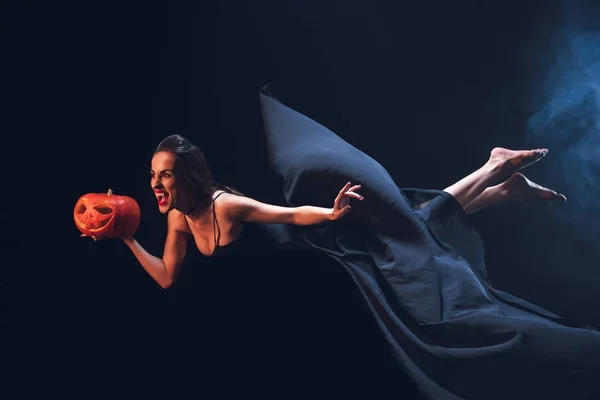 Donna in costume da vampiro in possesso di jack o lanterna e volare sul buio con il fumo — Foto stock