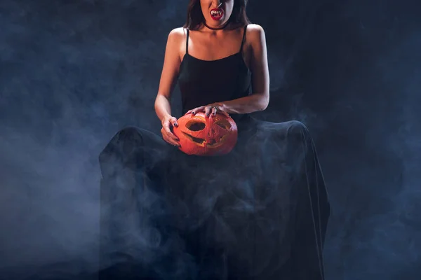 Cropped view of woman in vampire costume holding jack o lantern on darkness with smoke — Stock Photo