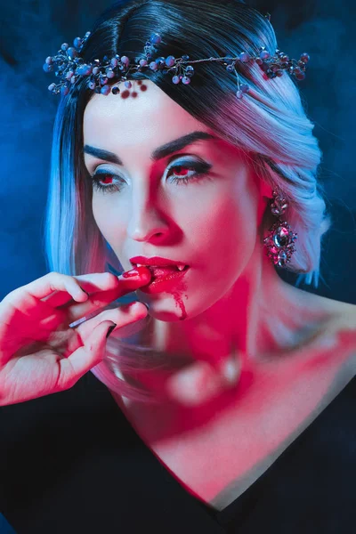 Sexy vampire woman licking bloody fingers on darkness with smoke — Stock Photo