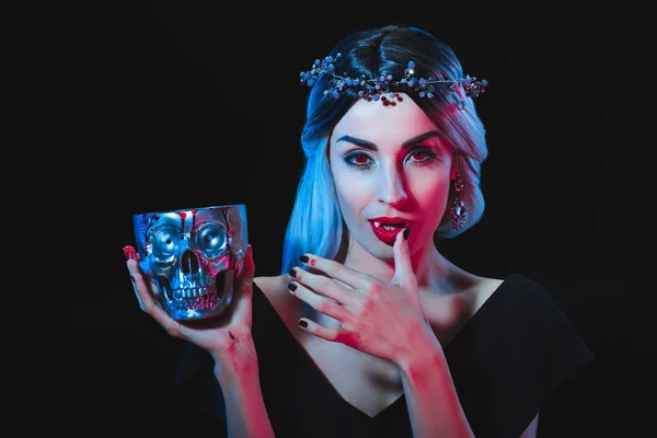 Vampire woman holding skull with blood and licking her fingers isolated on black — Stock Photo