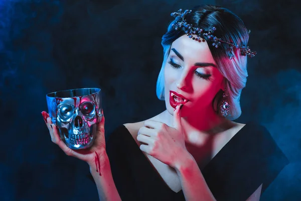 Beautiful vampire woman holding metal skull with blood and licking her fingers isolated on black — Stock Photo