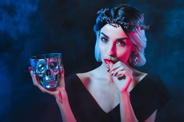 Sexy vampire woman holding skull with blood and licking her fingers isolated on black — Stock Photo