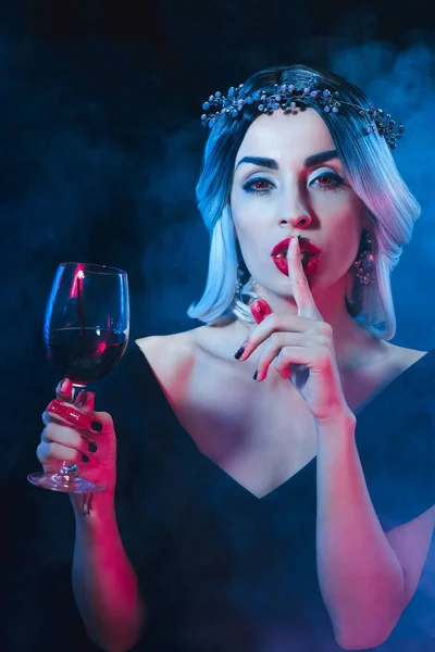 Sexy vampire woman showing silence symbol and holding wineglass with blood on dark background with smoke — Stock Photo