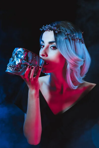 Sexy vampire woman drinking blood from metal skull on dark background with smoke — Stock Photo