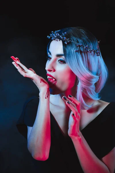 Attractive vampire licking blood from hand on dark background with smoke — Stock Photo