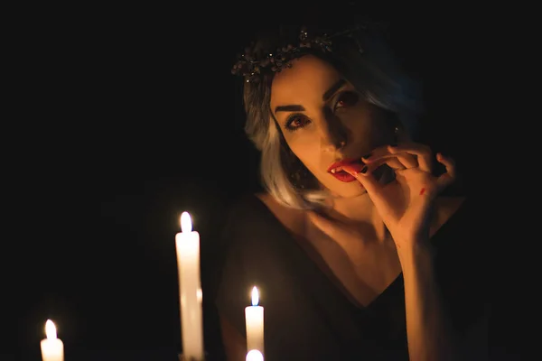 Beautiful woman in vampire costume licking her fingers with candles on foreground isolated on black — Stock Photo