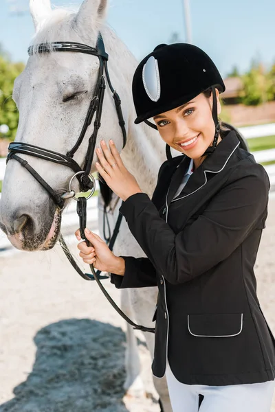 Atractiva hembra ecuestre en casco de montar mirando a la cámara cerca de caballo blanco en el club de caballos - foto de stock