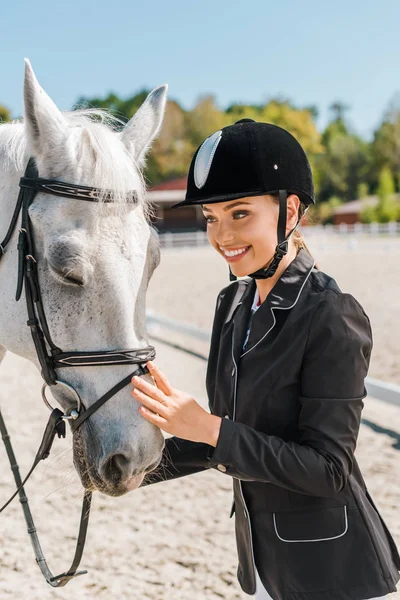 Atractivo sonriente hembra ecuestre de pie cerca de caballo en el club de caballos - foto de stock