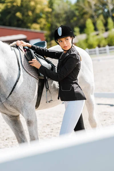 Красивая женщина конница фиксирует седло лошади в конном клубе — стоковое фото